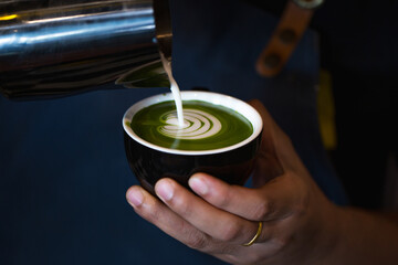 Close-up of a skilled hand crafting latte art in a green matcha latte. The image captures the moment milk meets tea, forming an elegant heart-shaped design.