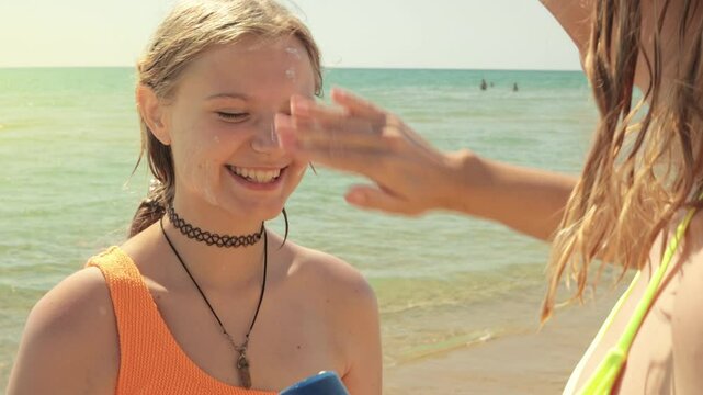Caring mother gently rubbing sunscreen onto teenager's face, shielding sensitive skin while relaxing near sparkling ocean waves during beach holiday.Active healthy lifestyle concept