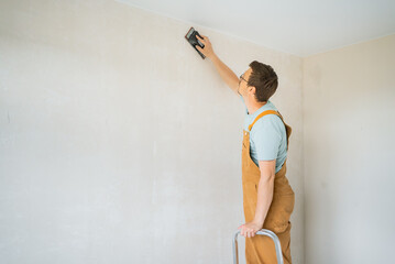 Construction worker wearing brown overalls, using an electric sander while standing on a metal...