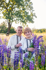 Smiling mature Ukrainian couple wearing vyshyvanka shirts and holding lupine flowers, standing...