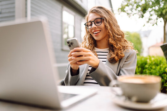 A young woman sits at a table on the veranda in a cafe with a laptop and a phone in her hands. A business woman drinks coffee and works with a laptop outdoors. Freelancing and coffee break concept.