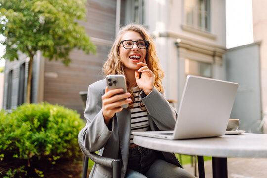 A young woman sits at a table on the veranda in a cafe with a laptop and a phone in her hands. A business woman drinks coffee and works with a laptop outdoors. Freelancing and coffee break concept.