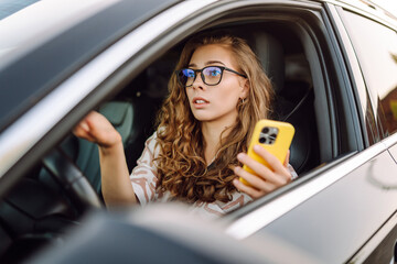 A young woman with curly hair driving a car and holding a phone in her hands on a sunny day. Woman...