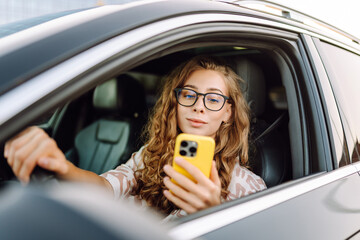 A young woman with curly hair driving a car and holding a phone in her hands on a sunny day. Woman...