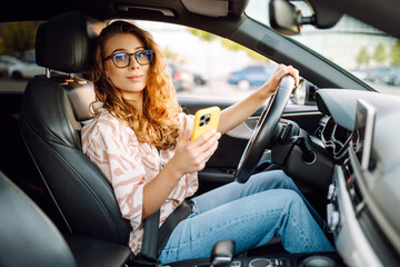 A young woman with curly hair driving a car and holding a phone in her hands on a sunny day. Woman...