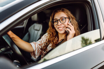 A young woman with curly hair driving a car and holding a phone in her hands on a sunny day. Woman...