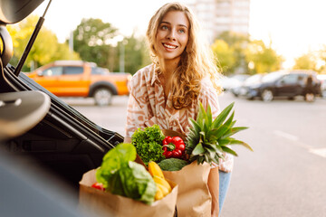 Smiling woman with bags full of fresh produce puts her purchases in trunk of her car at sunset. In...