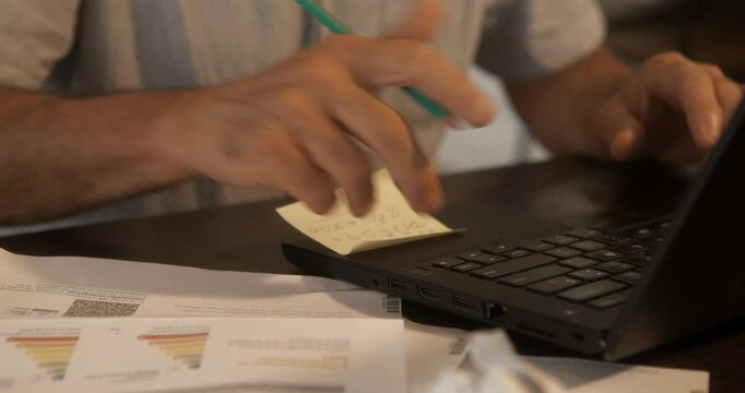 mature man's hands on the laptop keyboard checking invoices, pencil in hand, in his living room,
