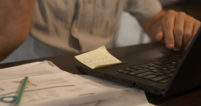 Man's hands on laptop keyboard checking invoices, with a pencil, in his living room. He has paperwork, suddenly throws the pencil over the invoices.