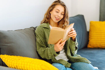 Young woman in glasses enjoying a book sitting on the sofa in a cozy room. Cute woman in glasses...