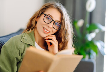 Young woman in glasses enjoying a book sitting on the sofa in a cozy room. Cute woman in glasses...