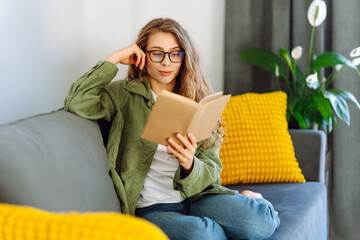 Young woman in glasses enjoying a book sitting on the sofa in a cozy room. Cute woman in glasses...