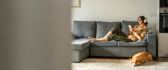 Smiling young student woman lying on couch, listening music in headphones on mobile phone...