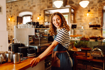 Happy female barista holding a cup of hot and aromatic coffee in her hands standing behind the bar....