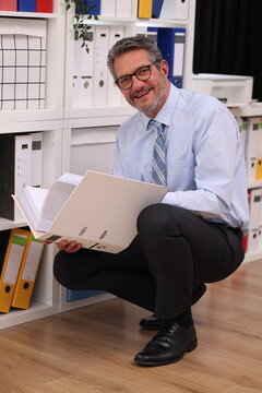 Office worker in shirt and tie kneels in front of a filing cabinet, holds file folders and smiles