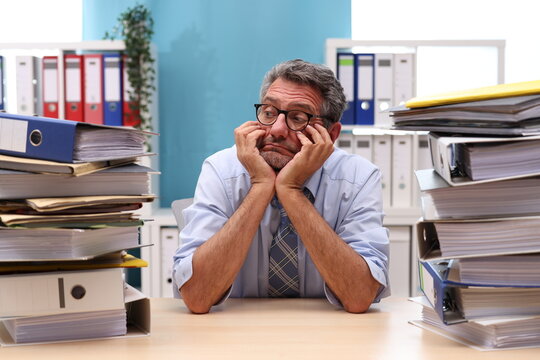 Office worker with glasses sits at the desk, supports his head with his arms and looks depressed to the side