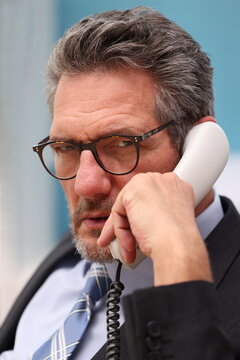 Office worker with glasses and suit sitting at his desk, talking on the phone and is very dissatisfied with the conversation, looking sternly ahead