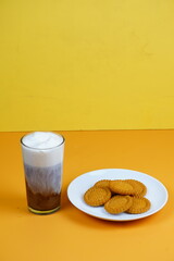 A refreshing glass of iced coffee with a foamy top and a plate of golden-brown biscuits, all against a vibrant orange backdrop.