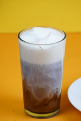 A close-up of a tall glass of iced coffee with a creamy white foam top, showing distinct layers of brown and white liquid against a bright orange background.