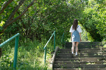 A young woman is ascending stone steps lined with a green railing, surrounded by vibrant foliage on a sunny day. The scene showcases the tranquility of nature and the joy of exploration