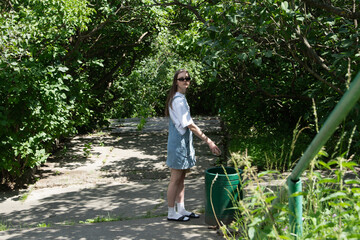 A woman stands outdoors in a green, overgrown pathway. She is wearing sunglasses and actively picking up litter while surrounded by dense foliage
