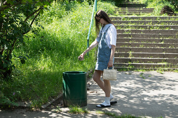 A young woman wearing a denim dress and sandals is throwing trash into a green bin while standing on a grassy path. Sunlight shines down on the scene, creating a bright atmosphere