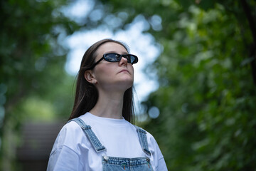 A young woman wearing sunglasses and a white shirt stands confidently in a park. Her long hair is flowing as she appreciates the calm atmosphere and natural beauty around her