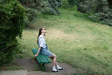 A woman sits casually on a green bench in a peaceful park. She gazes thoughtfully towards the sky, basking in the warm sunlight, with tall trees in the background and freshly cut grass nearby