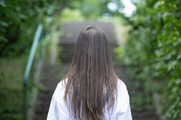 A person with long hair is seen from behind, ascending a set of stairs. Lush greenery lines the path, creating a serene atmosphere on a bright day, inviting contemplation