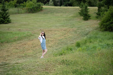 A girl stands in a lush green field, wearing sunglasses and a denim dress. She smiles playfully while enjoying the warm weather and the beautiful outdoor landscape