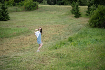 A young woman stands playfully in a lush green field, wearing casual attire and sunglasses. She smiles and poses, enjoying a beautiful day outdoors surrounded by nature