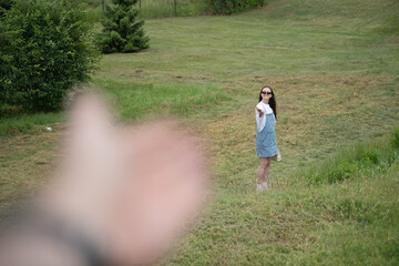 A young woman in a light dress stands in a lush green field, wearing sunglasses and smiling as she gestures. Bright daylight enhances the vibrant colors of the landscape
