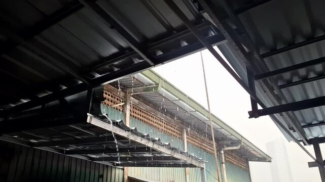 The side of a house with monsoon rains falling from its roof and hitting the roof of another house. The roof of the house is rusted and the gutters of the pipes that channel the water to the ground.