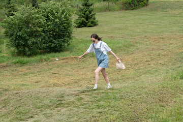 A young woman wearing denim overalls and sunglasses enjoys a warm, sunny day as she walks through a lush, grassy field. She carries a small bag while exploring the outdoors