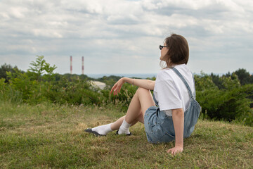 A young woman sits comfortably on a grassy hill, enjoying a moment of relaxation while gazing at the horizon. The cloudy sky adds a serene touch to the industrial view