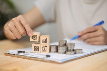 A hand places a block with a target icon onto a stack of other financial-themed blocks, symbolizing setting goals for financial growth and investment.