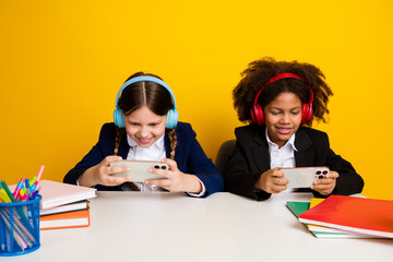Two cheerful schoolgirls play games together on phones, wearing headphones and sitting at a desk...