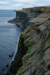 Dramatic coastal cliffs with a waterfall flowing into the sea in Iceland, showcasing rugged natural beauty, steep rock formations, and seabirds flying near the shoreline on a cloudy day