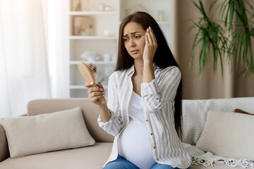 Worried pregnant woman at home looking at hairbrush with fallen hair, concerned about hair loss...
