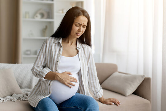 Pregnancy Pain. Young Pregnant Woman Touching Belly While Sitting On Couch At Home, Worried Expectant Female Suffering Painful Contractions Or Braxton Hicks, Making Breathing Exercises To Relief Ache