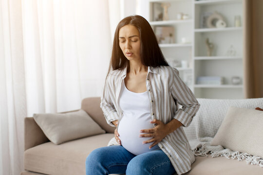 Braxton Hicks contractions. Young pregnant woman in pain suffering from abdominal ache at home, worried female sitting on couch, touching her big tummy, having cramp, copy space