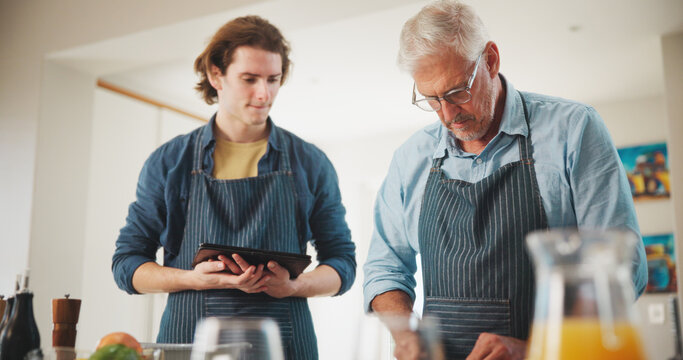 Mature man, chef and cooking with apprentice in kitchen for culinary skill or hospitality in home. Male people, mentor and helping junior with technology for breakfast, house recipe or food cuisine