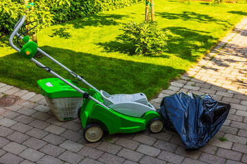 Electric lawn mower with grass bag and full black trash bag on stone pavement near green lawn. Sweden.