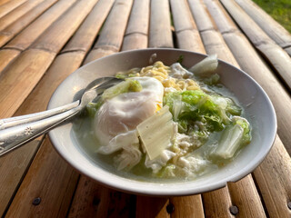 Hand movements holding spoons and forks, stirring a bowl of delicious steaming instant noodles, complemented by fried eggs and chopped green vegetables such as cabbage. The bowl is placed on a bamboo 