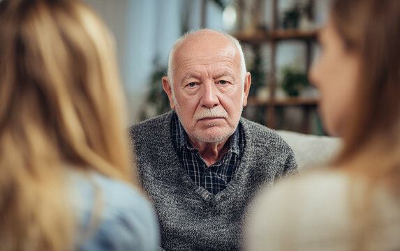 Elderly man sits in center speaking with two young women during home group therapy, photo captured from behind to highlight emotional conversation and supportive atmosphere.