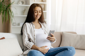 Happy pregnant millennial woman using smartphone at home, browsing pregnancy app while sitting on couch in living room, smiling expectant lady looking at phone screen and touching belly, free space
