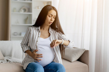 Pregnant woman checking time on her smartwatch while holding her belly, appearing to time...
