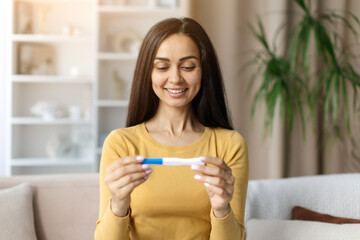 Happy young woman in yellow top smiling as she looks at a pregnancy test, sitting at home and...
