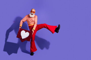 Energetic elder man in stylish retro outfit posing with a heart styled box against a vibrant purple background