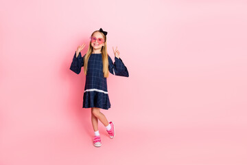 Charming young girl in stylish blue dress poses with peace signs on pink background, expressing...
